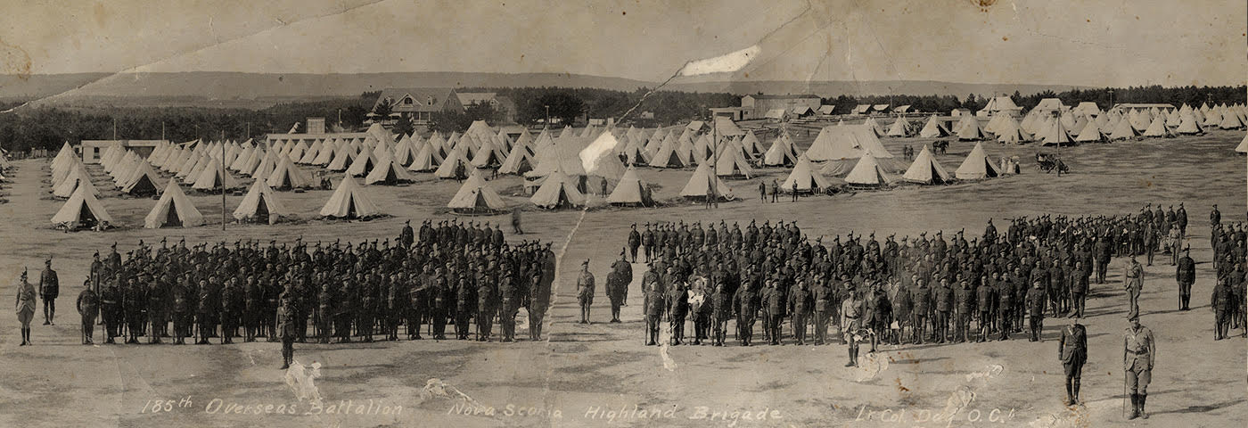 Photographie panoramique sépia de centaines de soldats en uniformes à un camp d’entraînement, avec le commandant à l’avant devant des rangées de tentes militaires en toile de fond.