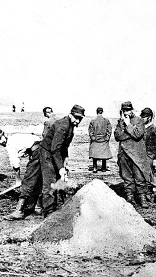 Item is a black and white photograph of a prisoner of war camp in Germany during the First World War. Several dozen men in uniform are pictured with shovels performing manual labour.
