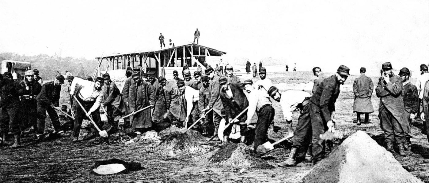 Il s’agit d’une photographie en noir et blanc d’un camp de prisonniers de guerre en Allemagne durant la Première Guerre mondiale. On voit des douzaines d’hommes en uniforme en train d’effectuer des travaux manuels à la pelle.