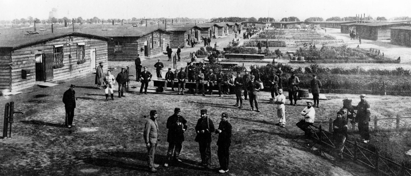 Black and white image of a prisoner of war camp, featuring groups of men standing outside, surrounded by military structures and gardens.