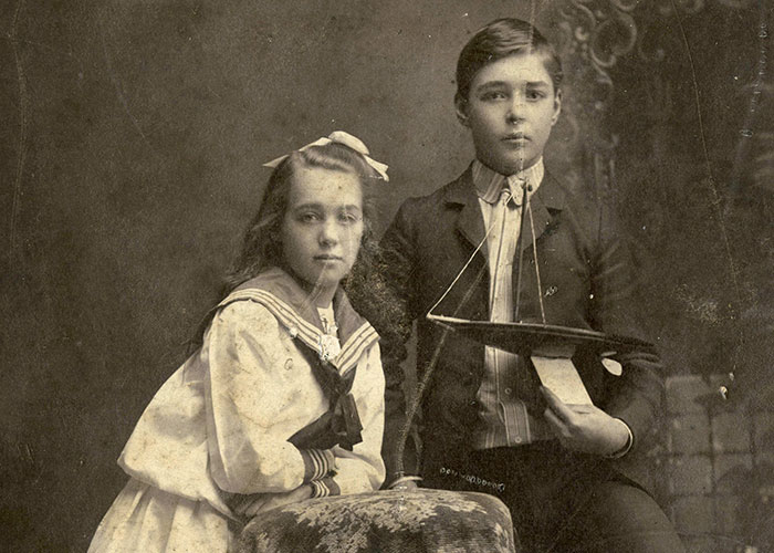 Black and white studio portrait of Helen Mary Kendall and her brother, Ernest James Kendall, as young children. Helen is on the left, wearing a white sailor dress with a bow in her hair. She is leaning on a covered stool. Ernest is standing on the right, holding a boat with his foot propped up on a fur rug.