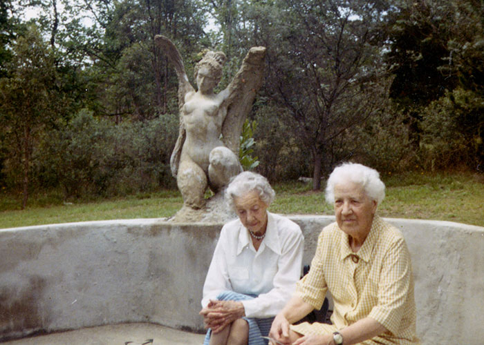 Colour photograph of two elderly women sitting outdoors in front of a fountain featuring an angel statue.