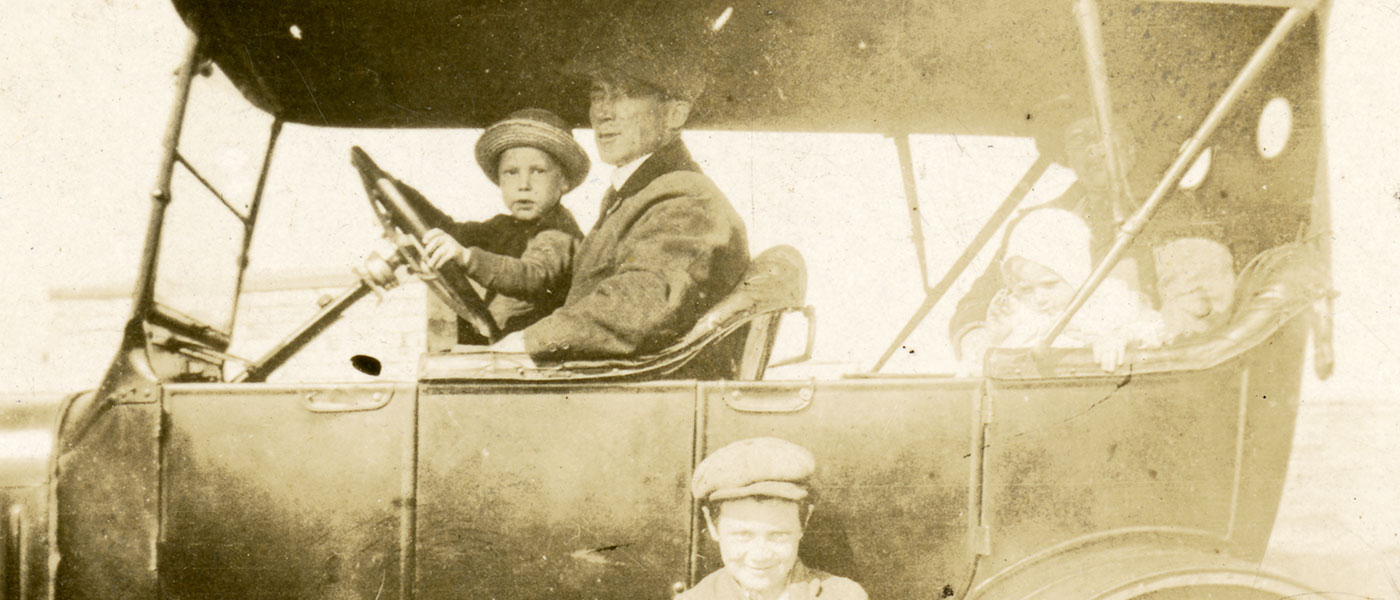 Sepia photograph of an old car. A man is posed at the drivers wheel with a child on his lap. There is a woman and a small child in the back of the automobile. A young boy smiles for the camera while standing outside of the car by its drivers door.