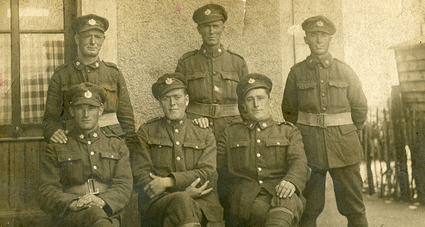Sepia photograph of six military men in uniform. Three men are sitting on a bench outdoors with their legs crossed. Three men are standing behind them.