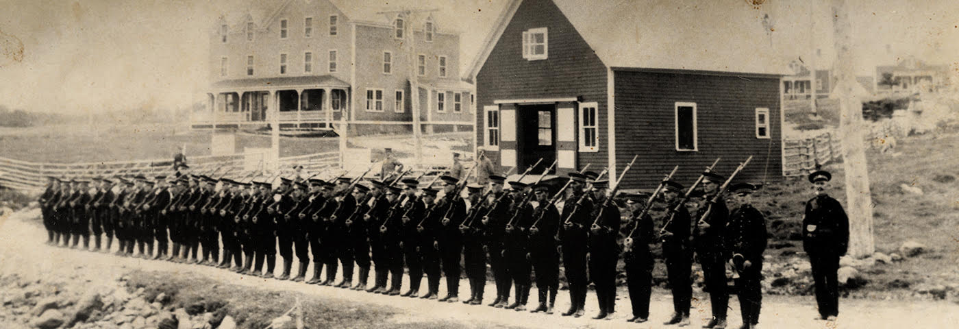 Black and white photograph of soldiers at arms standing on a dirt road in rural Cape Breton Island. There are two buildings in the distance, one of which is Marconi Station.