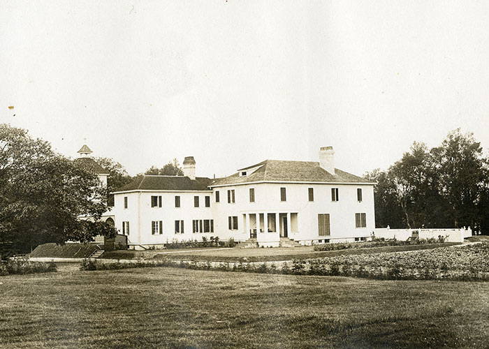 Photographie sépia d’une grosse maison blanche avec un grand chêne du côté gauche de la photo. La maison a deux cheminées.
