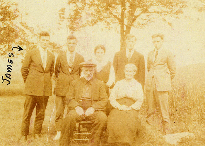 Photographie sépia d’une famille prise à l’extérieur. Les deux parents âgés sont assis sur des chaises à l’avant-plan avec cinq enfants debout derrière eux. Quelqu’un a écrit « James » à l’encre noire à côté de James Murphy debout à gauche dans la photo.